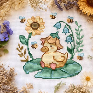 May include: Cross-stitch embroidery of a yellow duckling with a pink bow, on a green lily pad. The design includes a sunflower, bluebell flowers, and bees. The background is white, with dried flowers around the edges.