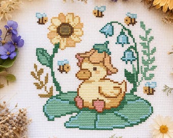 May include: Cross-stitch embroidery featuring a yellow duckling with a pink bow, sitting on a green lily pad. The design includes a sunflower, bluebell flowers, and bees. The background is white, with dried flowers around the edges.