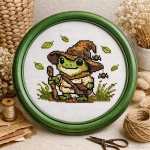 Può includere: Un ricamo a punto croce incorniciato raffigurante una rana verde con un cappello da mago marrone e un bastone. La rana è circondata da erba e foglie verdi, su sfondo bianco. La cornice è rotonda e verde scuro.