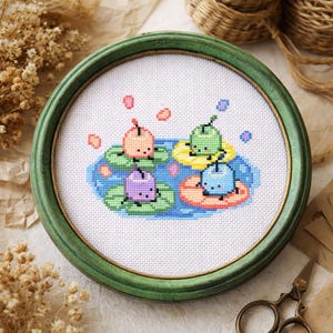 May include: A cross-stitch embroidery in a round, green frame. The design shows four cartoon characters on lily pads in a pond. The characters are in pastel colours, with pink, green, purple, and blue hues. The background is white.
