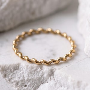 Könnte beinhalten: Ein dünner, goldfarbener Ring mit einem durchgehenden, sich wiederholenden gedrehten oder wellenförmigen Drahtdesign. Das Band weist eine glatte, polierte metallische Oberfläche auf, die das Licht reflektiert und ein gleichmäßiges Muster erzeugt.