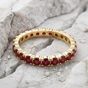 Könnte beinhalten: Ein goldener Eternity-Ring mit einer durchgehenden Reihe runder, tiefroter Rubine, die in einen polierten Gelbgoldrahmen gefasst sind. Der Ring ist kreisförmig und weist einen einheitlichen Fassungsstil auf, der leuchtend rote Edelsteine zeigt.