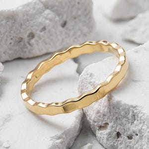 Può includere: Un anello in oro con un design a fascia ondulata continua. L'anello presenta una finitura metallica liscia e lucida che riflette la luce lungo i bordi curvi, creando un motivo costante di picchi e valli lungo l'intera circonferenza.