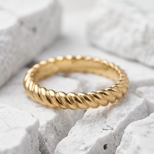 Könnte beinhalten: Ein goldener Ring mit einem durchgehenden gedrehten Seildesign. Das Band hat eine polierte, reflektierende Oberfläche mit einem sich wiederholenden diagonalen Muster, das ein strukturiertes Aussehen um den gesamten Umfang des Schmuckstücks erzeugt.