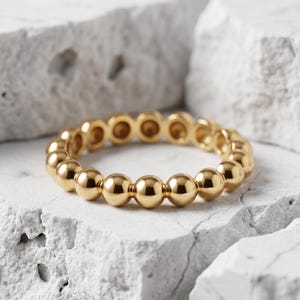 Puede incluir: Un anillo de tono dorado compuesto por una serie continua de pequeñas cuentas esféricas pulidas dispuestas en una banda circular. La superficie metálica refleja la luz, resaltando la forma uniforme y redondeada de cada cuenta del diseño.
