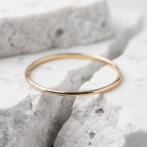 Può includere: Un sottile anello a fascia in oro lucido con una superficie liscia e arrotondata. L'anello presenta un design circolare semplice con una finitura metallica riflettente, mostrando un aspetto pulito e uniforme lungo tutta la circonferenza del gioiello.
