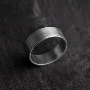 Könnte beinhalten: Ein gebürsteter Silberring mit flacher Oberseite und glattem, abgerundetem Innenraum. Der Ring ist vor einem dunklen, strukturierten Hintergrund platziert, der seinen metallischen Glanz hervorhebt. Dieses Schmuckstück ist ein schlichtes, modernes Design.
