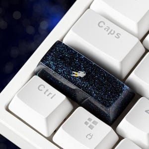 Può includere: Un tasto personalizzato per tastiera meccanica con un design spaziale blu navy profondo e scintillante, che presenta una minuscola figura di astronauta bianca al centro, posizionato tra tasti bianchi standard etichettati Ctrl, Caps e A.