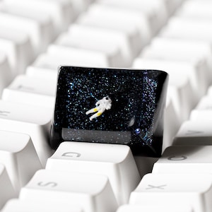 Può includere: Un tasto personalizzato per tastiera meccanica caratterizzato da uno sfondo nero profondo e scintillante a tema spaziale, con una minuscola e dettagliata statuetta di astronauta bianca e gialla incastonata al centro tra tasti bianchi.