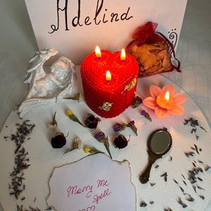 Op de afbeelding: Een rode hartvormige kaars met drie vlammen, gouden details en een getextureerd oppervlak. De afbeelding bevat een kleine spiegel, een wit engelenbeeldje en een papier met de tekst "Marry Me Spell". Het woord "Adelind" staat op een wit canvas.
