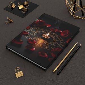 Könnte beinhalten: Ein schwarzes Notizbuch mit goldenem Design, einer brennenden Kerze und verstreuten roten Rosenblättern. Zwei Bleistifte und goldene Büroklammern sind ebenfalls vorhanden. Das Cover des Journals zeigt ein kreisförmiges Design mit Symbolen.
