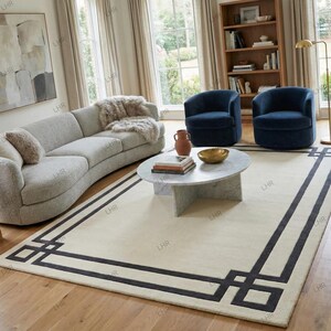 May include: A stylish living room with a light grey curved sofa, two navy blue armchairs, and a marble coffee table. A cream rug with a dark grey geometric border is the focal point. A bookshelf and large windows complete the room's decor.