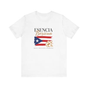 May include: A white short-sleeved t-shirt featuring a graphic design with the text ESENCIA Boricua, a Puerto Rican flag, and a gold hibiscus flower. Below the flag, the text reads MI ISLA • MI CULTURA • MI ORGULLO in a simple black font.