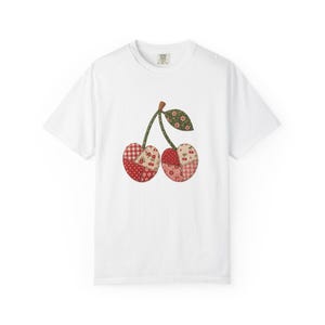 Puede incluir: Una camiseta blanca de manga corta con un gráfico central de dos cerezas. Las cerezas están diseñadas con un patrón de patchwork que consiste en varios estampados de tela rojos, blancos y verdes con motivos florales, de cuadros y lunares.