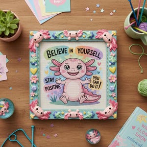 May include: A framed cross-stitch featuring a smiling pink axolotl with the text "Believe in Yourself, Stay Positive, You Can Do It!" The frame is light blue with axolotl and floral embellishments. The image is surrounded by craft supplies.