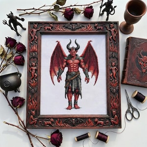 Può includere: Un'opera d'arte a punto croce incorniciata raffigura un demone rosso con grandi ali e corna. La cornice è intagliata con figure demoniache e fiamme. Rose secche, un calderone e altri oggetti gotici circondano l'opera.