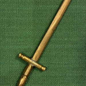 May include: A small, gold-colored sword with a cross-shaped hilt and a pointed blade. The sword is set against a textured green background. The sword's handle has a detailed, ornate design.