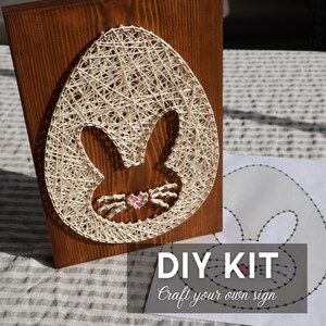 Op de afbeelding: Een houten paasei-vormig draadkunstwerk met een konijnensilhouet. De draad is gebroken wit en het hout is bruin gebeitst. Een roze hart zit in de neus van het konijn. De woorden "DIY KIT" en "Craft your own sign" zijn op papier gedrukt.
