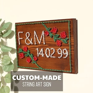 Puede incluir: Un letrero de arte de hilo hecho a medida sobre una tabla de madera. El letrero presenta las iniciales "F&M" y la fecha "14.02.99" en hilo blanco. Rosas rojas y hojas verdes de hilo también forman parte del diseño.