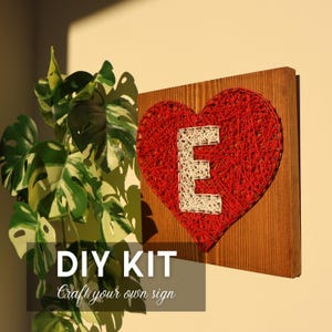 Puede incluir: Un letrero de madera con un corazón rojo hecho con arte de hilo, con la letra blanca "E" en el centro. El letrero está montado en una tabla de madera. El texto "DIY KIT Craft your own sign" está en la parte inferior.