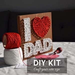 May include: DIY kit to create a string art sign that says "I ❤️ DAD". The wooden board features the words "I" and "DAD" in white string, and a red heart. Includes red and white string, and a tool. The kit also includes a paper that says "DIY KIT Craft your own sign".
