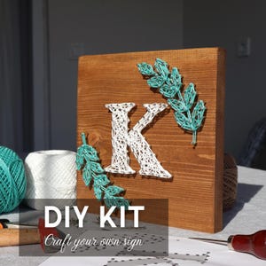 May include: Wooden sign with the letter "K" crafted with white string, adorned with teal string leaves. The sign is part of a DIY kit, with the text "DIY KIT Craft your own sign" at the bottom.