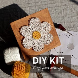 Op de afbeelding: Een DIY-set voor draadkunst met een houten plank met een wit en geel bloemmotief gemaakt met spijkers en draad. Bevat een bol wit garen, een bol geel garen, een papieren sjabloon, een hamer en een metalen priemgereedschap.