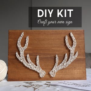 May include: DIY kit for crafting a sign. A wooden board with a dark stain features a string art design of deer antlers in white thread. The text "DIY KIT Craft your own sign" is at the top. A ball of string and small nails are also visible.