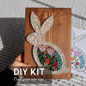 Op de afbeelding: Een doe-het-zelf-kit voor een draadkunstproject met een konijnontwerp. Het konijn is gemaakt van witte draad op een gebeitste houten plank, met roze en rode bloemen erin. De tekst "DIY KIT Craft your own sign" staat onderaan.