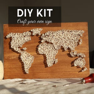Könnte beinhalten: DIY-Kit für eine String-Art-Weltkarte auf einer Holzplatte. Die Karte ist mit weißem Faden gefertigt und zeigt die Kontinente vor einem braunen Hintergrund. Der Text oben lautet "DIY KIT Craft your own sign."