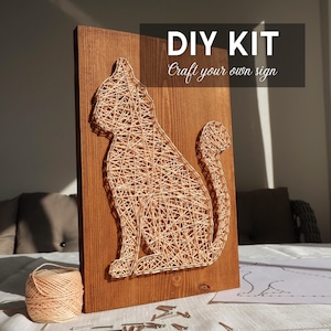 Puede incluir: Kit de bricolaje para un proyecto de arte de hilo de gato. El tablero de madera está teñido de marrón, con una silueta de gato hecha con hilo beige. El texto "DIY KIT Craft your own sign" se muestra en la parte superior. Incluye hilo, clavos y patrón.