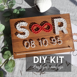 May include: A wooden string art sign with the letters "SOOR", an infinity symbol, and the numbers "08.10.95". The letters and numbers are white, the infinity symbol is red. The sign includes the text "DIY KIT Craft your own sign".