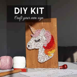 Könnte beinhalten: DIY-Kit für ein String-Art-Einhorn auf einer Holzplatte. Das Einhorn hat einen weißen Körper, eine rosa und rote Mähne und ein goldenes Horn. Enthält Faden, Nägel und Werkzeuge. Der Text "DIY KIT Craft your own sign" steht oben.