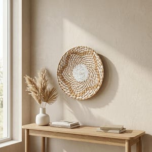 May include: A round, woven wall basket with a spiral design, featuring natural and white tones. The basket is mounted on a beige wall. A wooden console table with a white vase and books is in the foreground.
