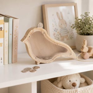 May include: A bird-shaped wooden piggy bank with a clear acrylic front panel, featuring a coin slot at the top. The natural light-colored wood frame is secured with small metal screws. Several metallic coins are visible inside and on the surface.