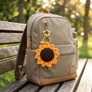 以下が含まれることがあります： カーキ色のキャンバスバックパック。茶色の革のアクセントと、かぎ針編みのヒマワリのキーホルダーが付いています。ヒマワリは茶色の中心とオレンジ色の花びらです。黄色の星とベルのチャームも付いています。
