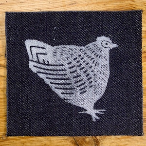 Könnte beinhalten: Ein quadratischer, dunkelblauer Denim-Patch mit einem weißen Hühnerdesign. Der Patch liegt auf einer Holzoberfläche neben einem Stück Denim-Stoff. Das Huhn blickt nach rechts, mit detaillierten Feder- und Flügelmustern.