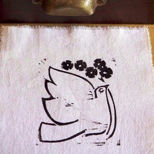 Könnte beinhalten: Ein weißes Leinentuch mit einem schwarzen Tauben- und Blumendesign. Die Taube ist stilisiert mit einem gebogenen Hals und Flügel, und die Blumen sind über ihrem Kopf angeordnet. Das Tuch hat einen rohen, unfertigen Rand.