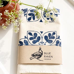 Könnte beinhalten: Ein gefaltetes cremefarbenes Textil mit einem dunkelblauen botanischen Blockdruckmuster. Ein beigefarbenes Papierband umschließt die Mitte und zeigt eine schwarze Strichzeichnung eines Vogels sowie den Text "BLUE RAVEN FOLKWEAR".