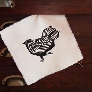 Könnte beinhalten: Ein quadratisches, cremefarbenes Stoffstück mit einem schwarzen Linolschnitt eines Huhns. Das Huhn ist im Profil dargestellt, mit detaillierten Federn und einem strukturierten Körper. Der Stoff hat einen rohen, unfertigen Rand und wirkt rustikal.