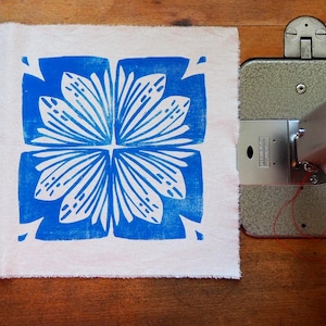 Op de afbeelding: Een vierkant witte stof met een blauw bloemenpatroon ligt naast een vintage naaimachine. Het blauwe ontwerp is een symmetrisch patroon van gestileerde bloemblaadjes. De naaimachine is zilverkleurig en heeft rode draad.