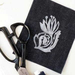 Könnte beinhalten: Ein quadratischer Aufnäher aus schwarzem Denim mit einem silbernen, gestickten Blumenmuster. Neben dem Aufnäher liegen eine schwarze Metallschere mit japanischen Schriftzeichen auf der Klinge und eine kleine Rolle dunkelblaues Nähgarn.