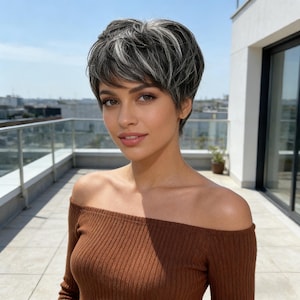 Könnte beinhalten: Kurze, gestufte Perücke mit grauen und weißen Strähnen. Die Perücke hat einen seitlich geschwungenen Pony und einen natürlichen Haaransatz. Die Person trägt ein braunes schulterfreies Oberteil. Der Hintergrund zeigt eine sonnige Umgebung im Freien.