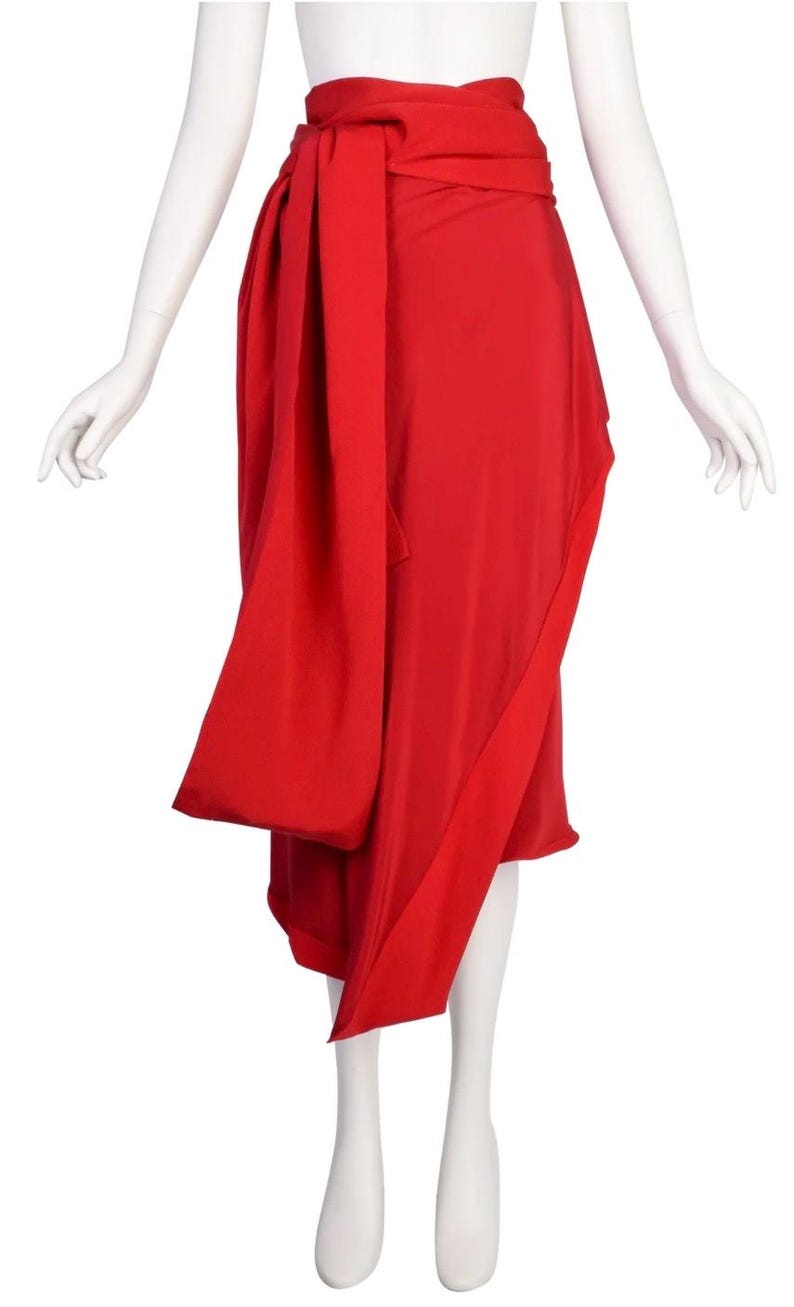 May include: A bright red skirt with a wrap-around design. The skirt has a long, flowing sash that wraps around the waist and hangs down the front. The fabric looks lightweight and has a slight sheen.