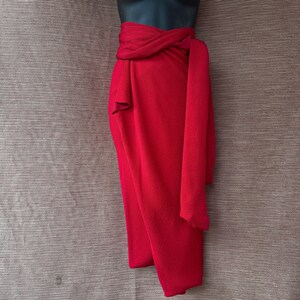 May include: A striking red wrap skirt, elegantly draped and tied at the waist. The fabric falls gracefully, forming soft folds and a flowing shape. The skirt is displayed on a black mannequin, highlighting its design and texture.