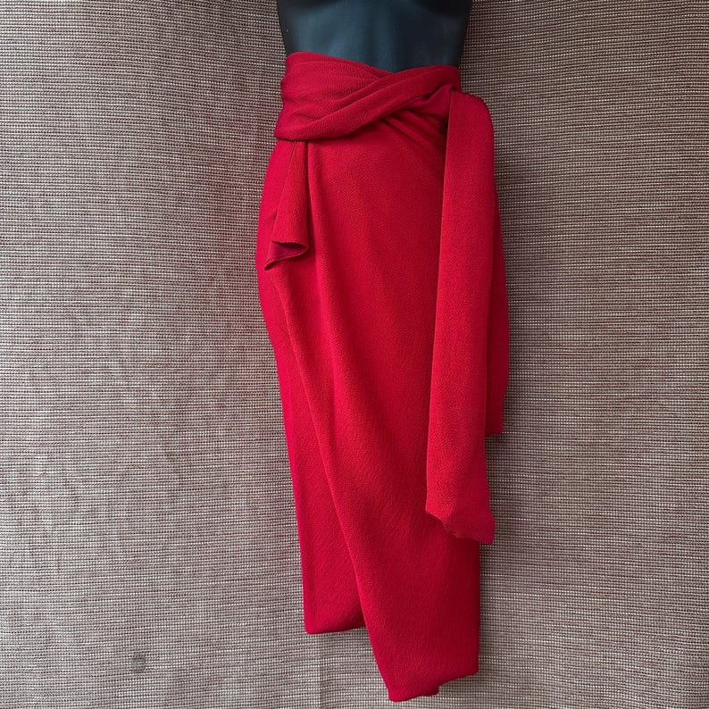 May include: A striking red wrap skirt, elegantly draped and tied at the waist. The fabric falls gracefully, forming soft folds and a flowing shape. The skirt is displayed on a black mannequin, highlighting its design and texture.