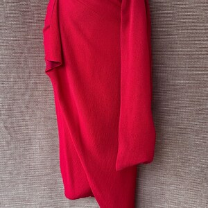 May include: A bright red wrap skirt with a draped design. The skirt is made of a textured fabric and features a wrap-around style with a long, flowing sash. The skirt is displayed on a mannequin.