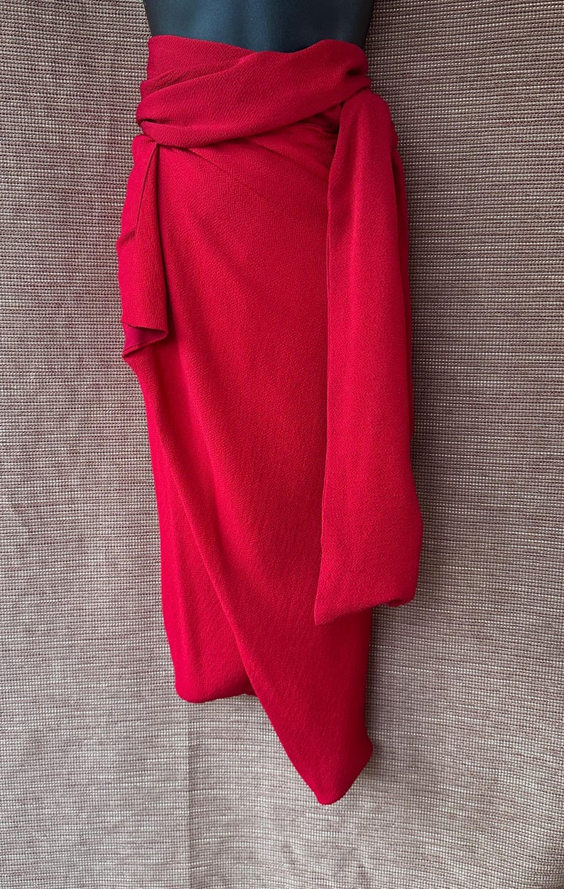 May include: A bright red wrap skirt with a draped design. The skirt is made of a textured fabric and features a wrap-around style with a long, flowing sash. The skirt is displayed on a mannequin.