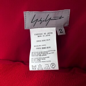 May include: A red garment with a label showing the designer's signature and care instructions. The label says "FABRIQUE AU JAPON MADE IN JAPAN" and "100% SOIE SILK". The garment is made of 100% silk.