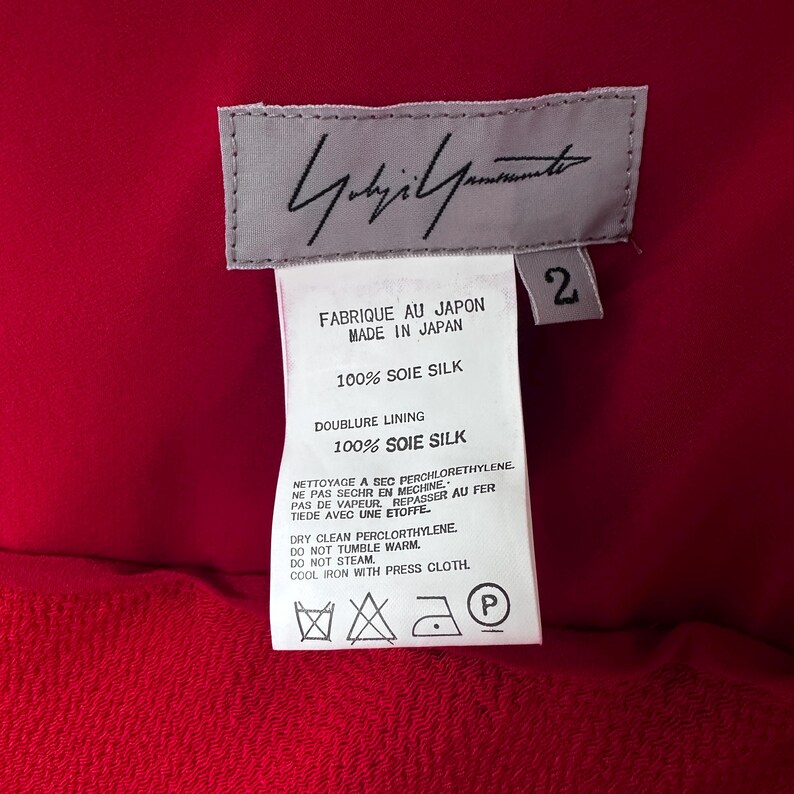 May include: A red garment with a label showing the designer's signature and care instructions. The label says "FABRIQUE AU JAPON MADE IN JAPAN" and "100% SOIE SILK". The garment is made of 100% silk.